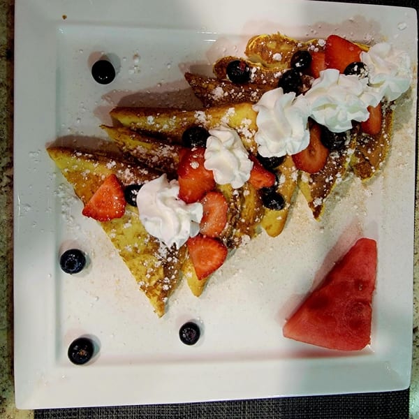 French toast triangles topped with whipped cream, strawberries, blueberries, and powdered sugar, with a watermelon slice.