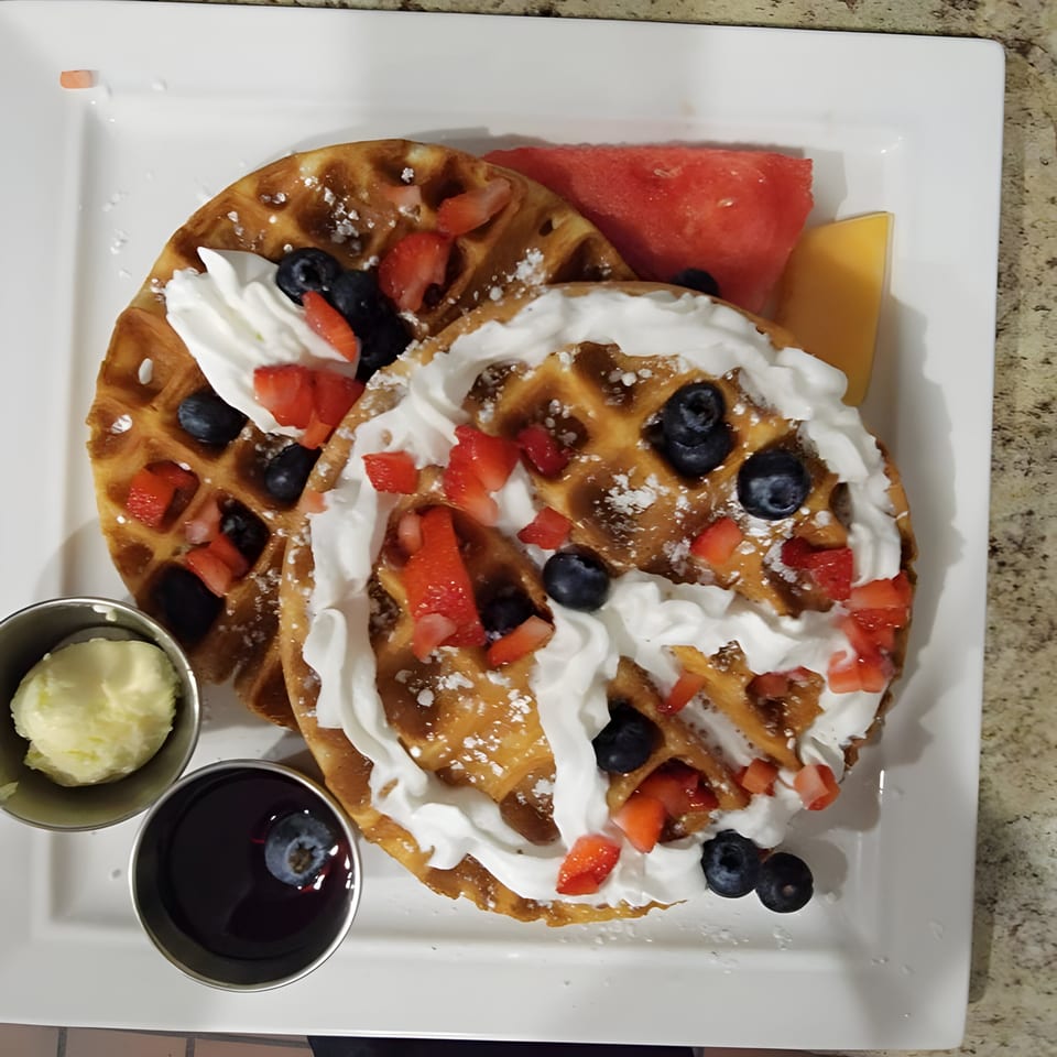 Two waffles topped with whipped cream, strawberries, blueberries, powdered sugar, and a side of fruit.