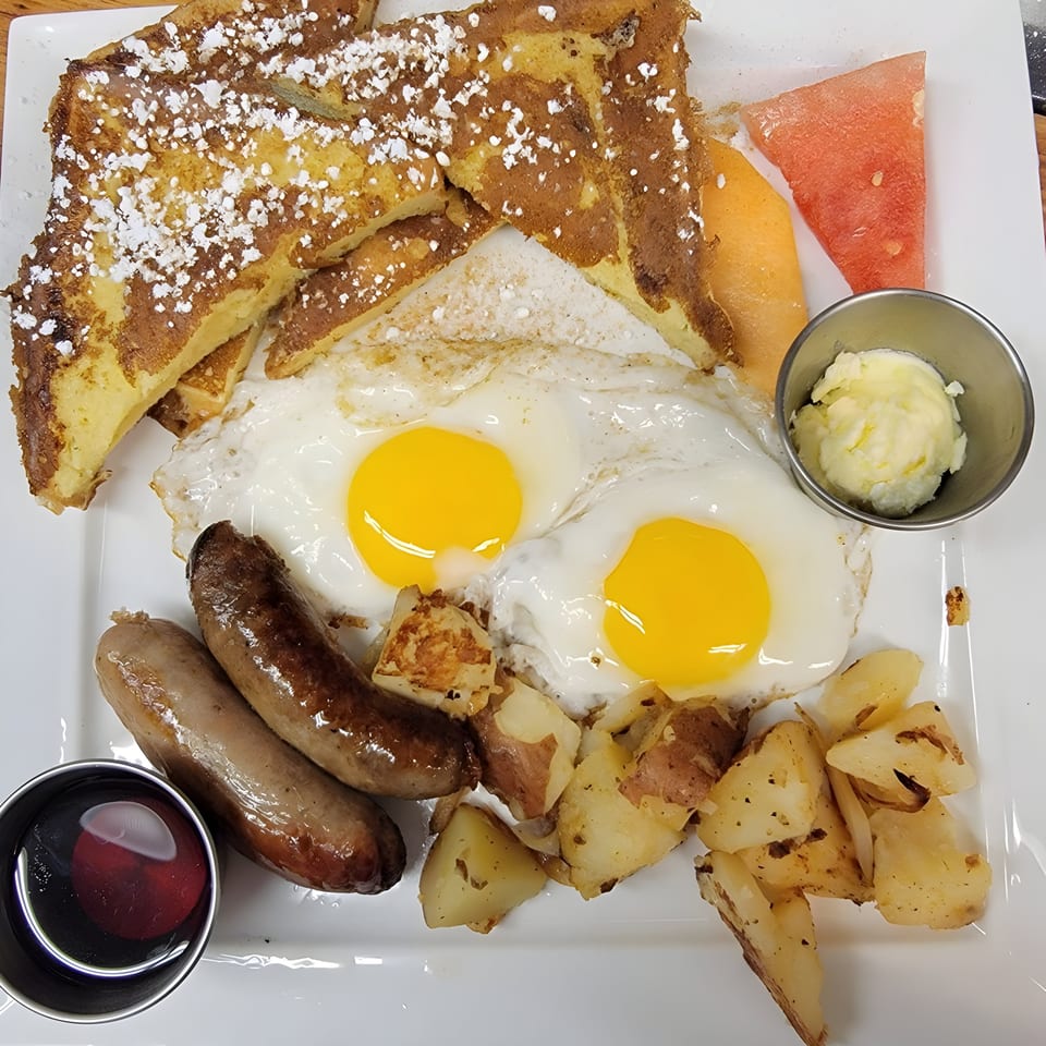 A hearty American breakfast with French toast, two fried eggs, sausages, home fries, and fruit.