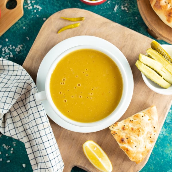 Overhead view of a bowl of yellow lentil soup, with pickles, lemon, and flatbread on a wooden board.
