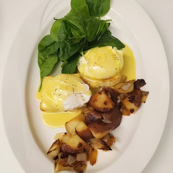 Eggs Benedict with hollandaise sauce, fresh spinach, and roasted potatoes on a white plate.