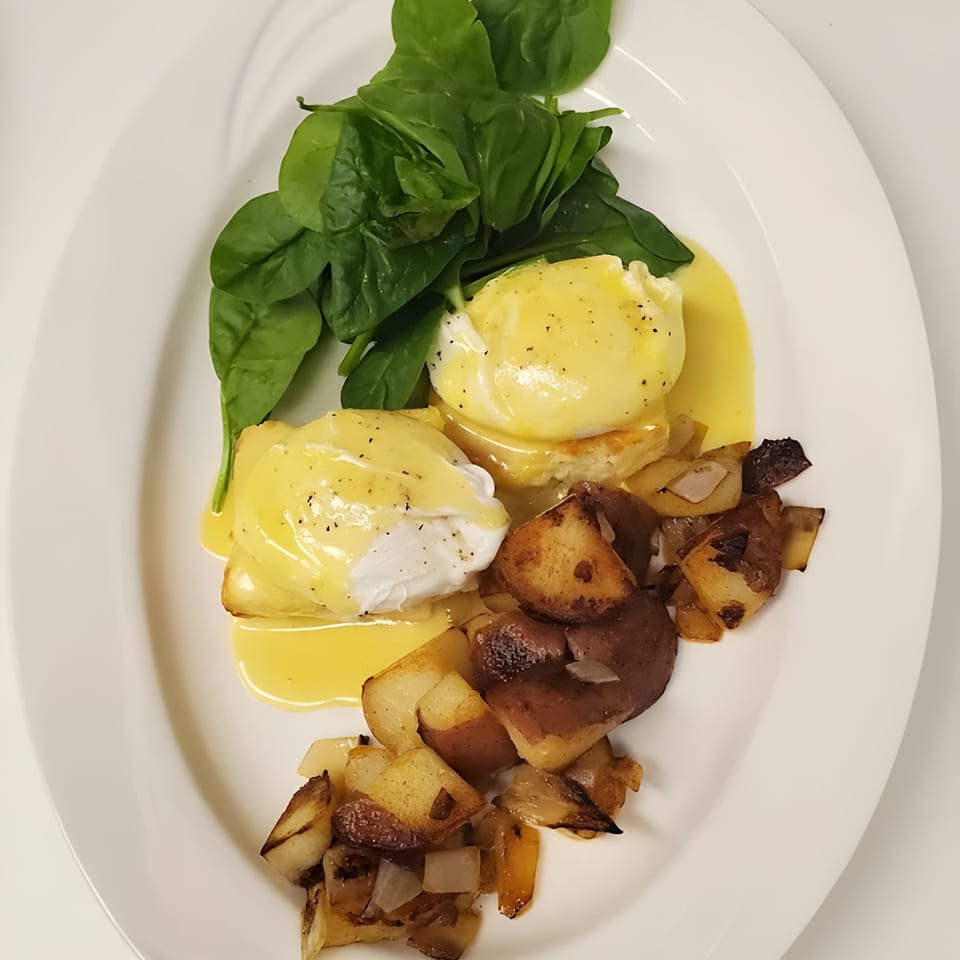 Eggs Benedict with hollandaise sauce, fresh spinach, and roasted potatoes on a white plate.